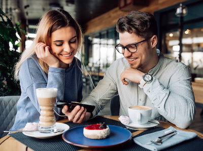 Blonde Frau und Mann sitzen mit Kaffee vor Tisch und schauen gemeinsam auf das Smartphone des Mannes
